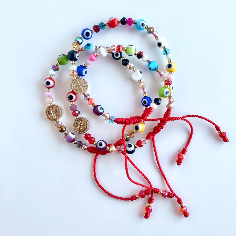May include: Three red string bracelets with evil eye charms and gold coin charms. The bracelets are layered on top of each other.