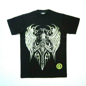 Peut inclure: T-shirt noir avec un imprimé graphique d'un crâne avec des ailes et une épée. Le crâne est entouré de motifs de style tribal. Le t-shirt a une petite étiquette verte et jaune avec le texte "Glow in the Dark".