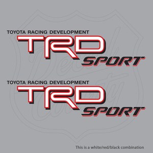 Puede incluir: Dos calcomanías TRD Sport. Cada una presenta el texto "TOYOTA RACING DEVELOPMENT" sobre las letras "TRD" en blanco con contorno rojo, y "SPORT" en negro. El fondo es gris con contorno blanco.