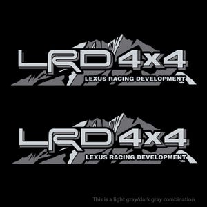 Op de afbeelding: Twee grijze en witte stickers met de tekst "LRD 4x4" en "LEXUS RACING DEVELOPMENT" op een zwarte achtergrond. De stickers tonen een gestileerde bergketen.