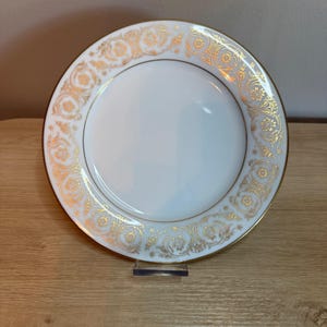 May include: A round white porcelain plate featuring a wide border decorated with an intricate gold filigree pattern of floral and scroll motifs. The rim of the plate is finished with a thin gold band, and the center remains plain white.