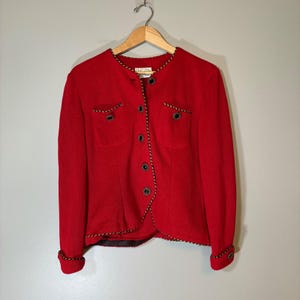 May include: A bright red long-sleeved jacket featuring a round neckline, a front button closure, and two chest pockets. The edges of the collar, front placket, pockets, and cuffs are trimmed with a decorative black and gold twisted rope piping.