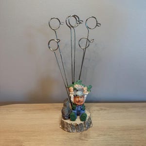 Può includere: Un supporto in metallo con tre sottili steli verticali in filo metallico terminanti con anelli circolari. La base è una statuetta dipinta di una persona barbuta con salopette blu e cappello, che tiene un pesce verde su un ceppo.