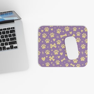 Peut inclure: Un tapis de souris rectangulaire avec un motif répétitif d'empreintes de pattes, d'os, de cœurs et de petites étoiles jaune clair sur un fond violet uni. Une souris d'ordinateur blanche repose sur le tapis à côté d'un ordinateur portable argenté.