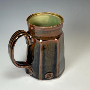 May include: A brown and black ceramic mug with a green glazed interior. The mug has a unique octagonal shape and a handle with a textured design. The mug is marked with a small circular logo on the bottom.