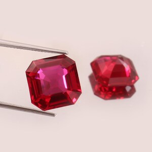 May include: Two faceted, rectangular-cut, deep red gemstones. One is held by tweezers, the other is reflected on a white surface. The gems have a vibrant, polished appearance, showcasing their internal structure and color.