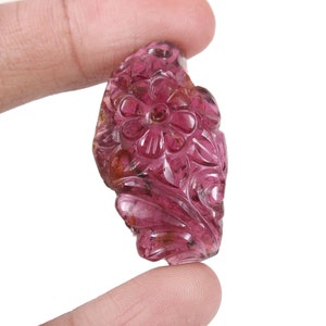 Puede incluir: Un colgante de piedra preciosa rosa tallada con un diseño floral. El colgante se sostiene entre los dedos, mostrando los intrincados detalles de los tallados de flores y hojas. La piedra preciosa es translúcida.