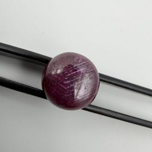 Pu&ograve; includere: Una pietra preziosa rotonda di colore magenta intenso con una superficie liscia e lucida e inclusioni cristalline interne visibili, tenuta saldamente tra le due sottili punte metalliche nere di una pinzetta su uno sfondo bianco.