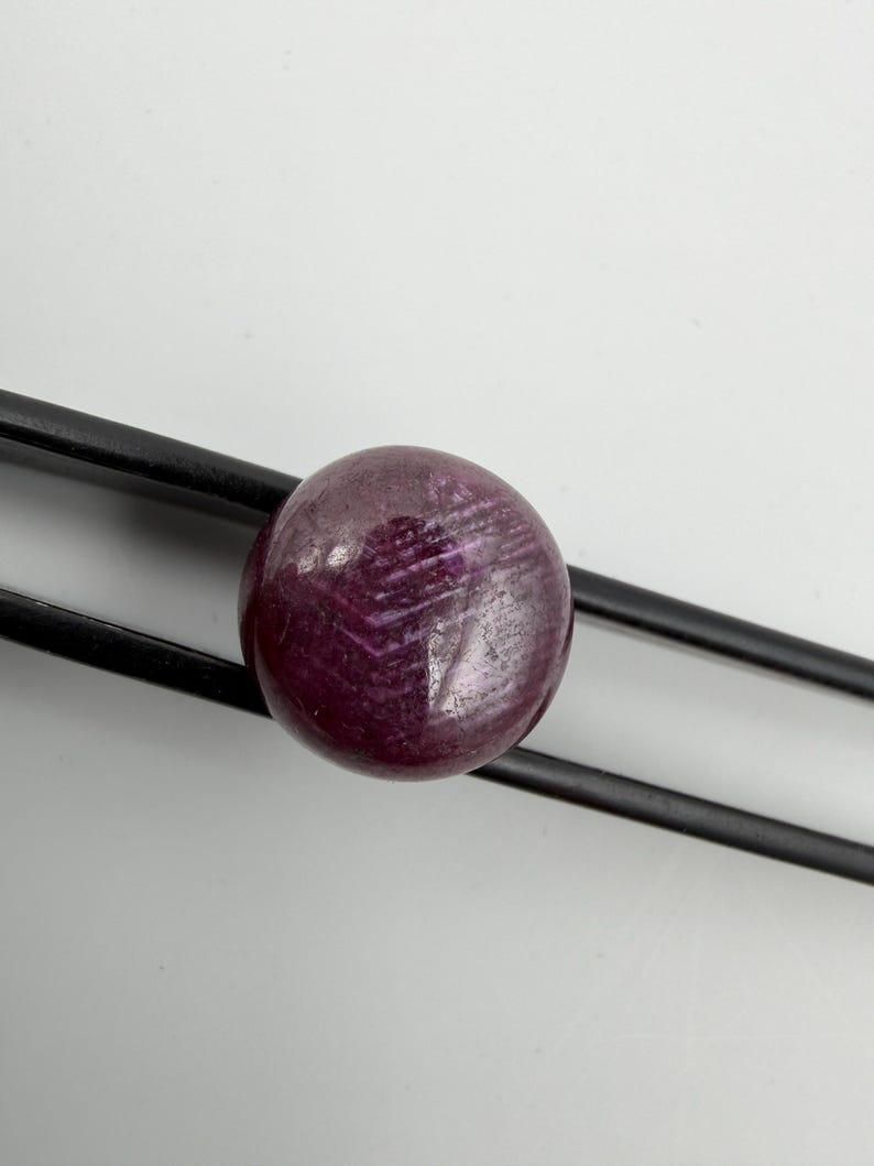 Pu&ograve; includere: Una pietra preziosa rotonda di colore magenta intenso con una superficie liscia e lucida e inclusioni cristalline interne visibili, tenuta saldamente tra le due sottili punte metalliche nere di una pinzetta su uno sfondo bianco.