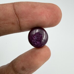 Pu&ograve; includere: Una piccola pietra preziosa rotonda tagliata a cabochon con una tonalit&agrave; profonda e satura di viola scuro e magenta. La superficie della pietra presenta una finitura liscia e lucida con striature interne e inclusioni naturali visibili.