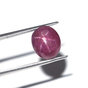Puede incluir: Una gema de rubí pulida, de forma ovalada, sujeta con pinzas sobre un fondo blanco. El rubí exhibe un efecto de estrella, con un patrón de estrella de seis rayos visible en su superficie. La gema es de un color rojo intenso.