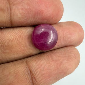 Könnte beinhalten: Ein runder, im Cabochon-Schliff gefertigter Edelstein in einer tiefen, gesättigten magentaroten Farbe. Der Stein hat eine glatte, polierte Oberfläche mit subtilen inneren Einschlüssen und einem reflektierenden Glanz, der ein lebendiges und opakes Erscheinungsbild bietet.