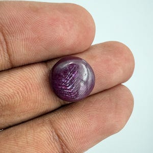 Puede incluir: Una piedra preciosa de rubí de corte cabujón redondo con un tono rojo violáceo profundo. La piedra muestra un fenómeno óptico en forma de estrella conocido como asterismo, con rayos de luz que se cruzan en su superficie pulida.