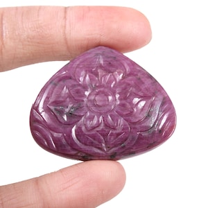 May include: A carved pink ruby gemstone with a floral design. The gemstone is shaped like a triangle with rounded edges.