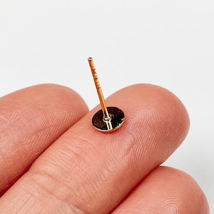Può includere: Un piccolo orecchino a perno color oro con una base circolare piatta e un perno sottile e dritto che si estende verticalmente dal centro. Il metallo presenta una superficie lucida e riflettente, con una calda tonalità giallo metallizzato.