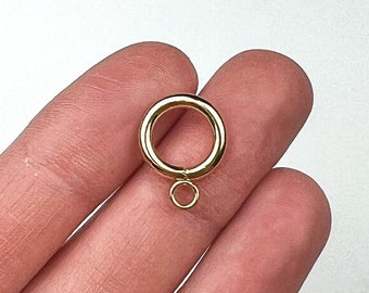 May include: A gold-coloured jewellery finding, featuring a large ring with a smaller ring attached below. The finding is held in a hand against a white background. The jewellery finding is likely used for connecting jewellery pieces.