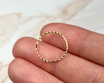 May include: A small, gold ring with a twisted rope design. The ring is held between fingers, highlighting its delicate details and reflective surface. The ring's design features alternating gold and dark lines.