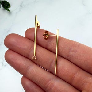 May include: A pair of gold-toned metal bar earrings. Each earring features a long, slender vertical bar design with a small circular loop at the bottom end and a post-style backing mechanism attached to the upper portion of the bar.