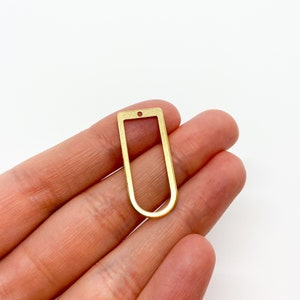 May include: A gold-toned metal pendant with a rectangular shape and a rounded top and bottom. The pendant has a small hole at the top for attaching a chain or cord.