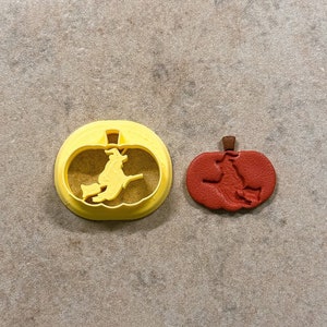 May include: Two Halloween-themed cookie cutters. One is yellow and shaped like a pumpkin with a witch flying on a broomstick inside. The other is red and shaped like a pumpkin with a witch flying on a broomstick inside.