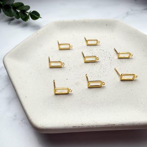 Pode incluir: Um conjunto de sete bases para brincos em metal dourado, cada uma com uma pequena moldura retangular, um pino e um pequeno elo na parte inferior para prender pendentes, dispostos sobre uma bandeja hexagonal de cor esbranqui&ccedil;ada.