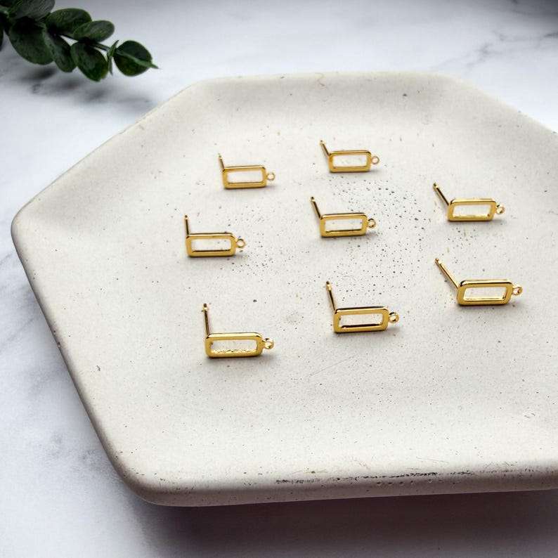 Pode incluir: Um conjunto de sete bases para brincos em metal dourado, cada uma com uma pequena moldura retangular, um pino e um pequeno elo na parte inferior para prender pendentes, dispostos sobre uma bandeja hexagonal de cor esbranqui&ccedil;ada.