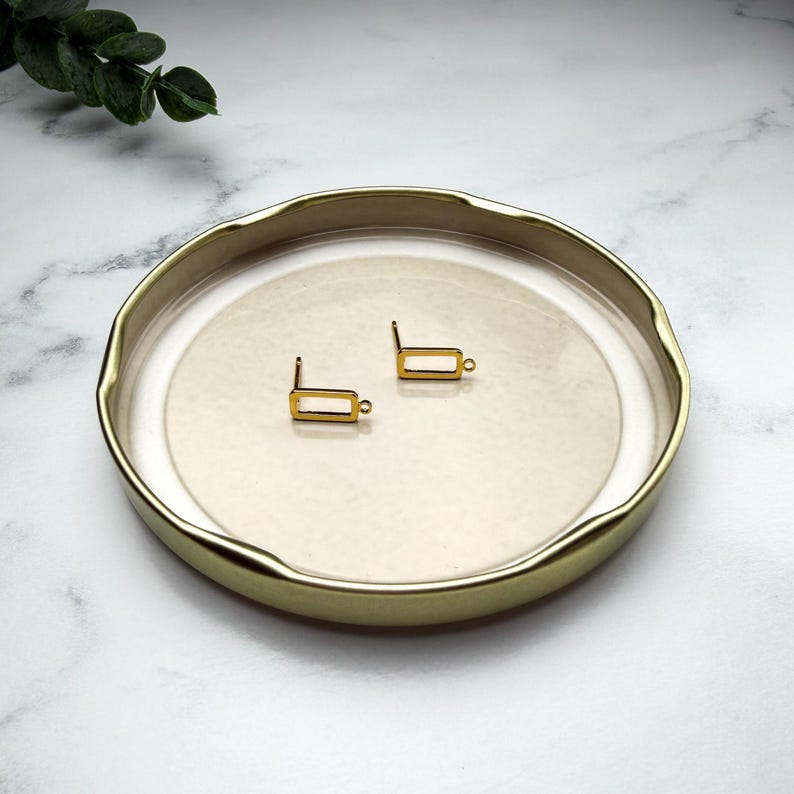Pode incluir: Um par de componentes de brinco em metal dourado, cada um apresentando uma pequena moldura retangular com um min&uacute;sculo elo circular preso a um lado, repousando sobre um prato redondo raso com borda dourada e interior liso.