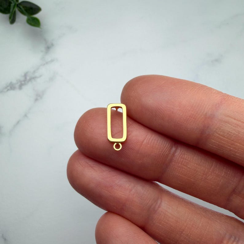 Pode incluir: Um componente de brinco em metal dourado com uma estrutura retangular oca e um pequeno anel de jun&ccedil;&atilde;o circular fixado na borda inferior, projetado para a cria&ccedil;&atilde;o de joias e montagem de acess&oacute;rios pendentes.