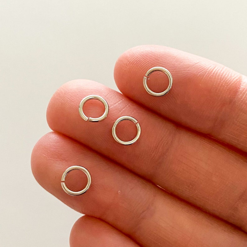 K&ouml;nnte beinhalten: F&uuml;nf silberne Sprungringe, auch bekannt als Splintringe, werden auf einer Hand gezeigt. Die Ringe sind klein und rund, mit einer L&uuml;cke im Kreis.