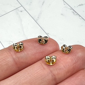 Peut inclure: Quatre dos d'oreille en or, également appelés bouchons d'oreille, sont présentés sur une main. Les dos d'oreille sont conçus pour maintenir les boucles d'oreilles en place.
