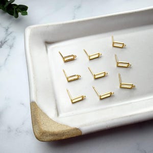Pode incluir: Um conjunto de oito pinos para brincos em metal dourado, cada um apresentando um design de moldura retangular pequena com um pequeno elo na parte inferior para prender pingentes. Estes componentes de joalharia est&atilde;o dispostos.
