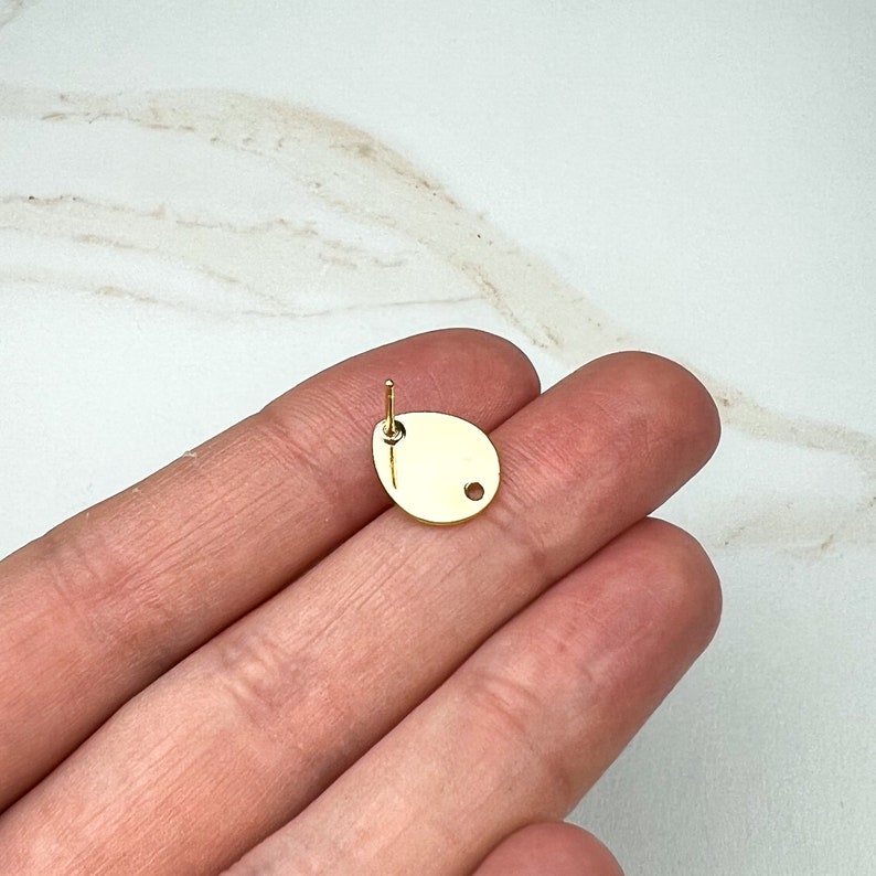 May include: Gold-toned metal earring finding with a round hole in the center. The finding is oval-shaped and has a post on the back.