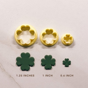 May include: Three clover-shaped cookie cutters and three dark green clovers. The cutters are yellow and round, with the clover shape cut out. The clovers are 1.25 inches, 1 inch, and 0.6 inches in size. The image is on a light gray surface.