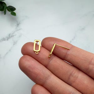 Pode incluir: Um par de brincos de pino dourados com um design de moldura retangular oca. Cada brinco tem um pino vertical fixado na parte de tr&aacute;s do ret&acirc;ngulo, com um pequeno elo circular na borda inferior para a fixa&ccedil;&atilde;o de componentes adicionais de joias.