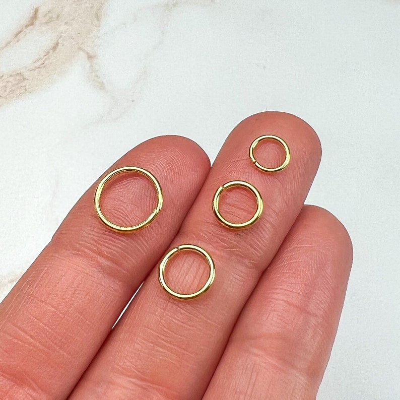 May include: Four gold-colored hoop earrings of different sizes. The hoops are displayed on a person's fingers.