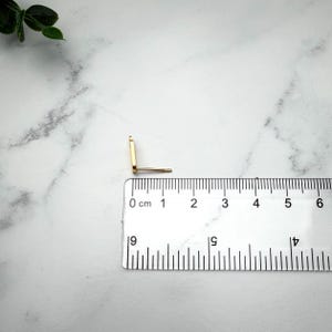 Pode incluir: Um brinco de pino dourado com uma face retangular e um pino, posicionado ao lado de uma r&eacute;gua de pl&aacute;stico transparente que mostra que o brinco mede aproximadamente 1,2 cm de comprimento da parte superior &agrave; inferior da face retangular.