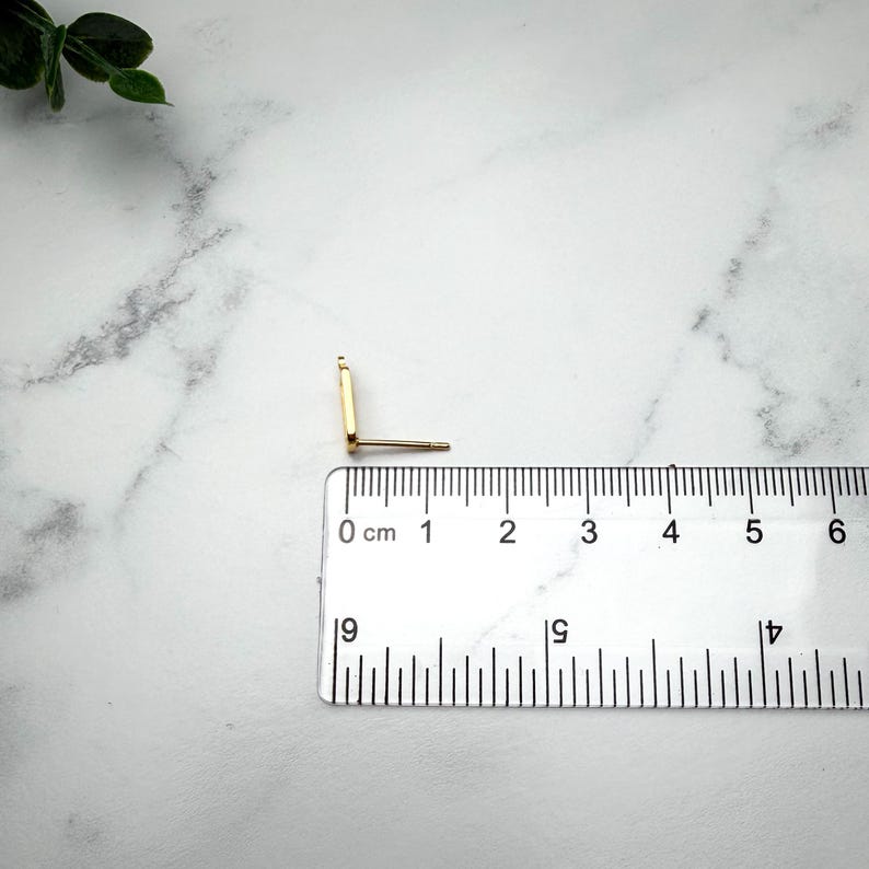 Pode incluir: Um brinco de pino dourado com uma face retangular e um pino, posicionado ao lado de uma r&eacute;gua de pl&aacute;stico transparente que mostra que o brinco mede aproximadamente 1,2 cm de comprimento da parte superior &agrave; inferior da face retangular.