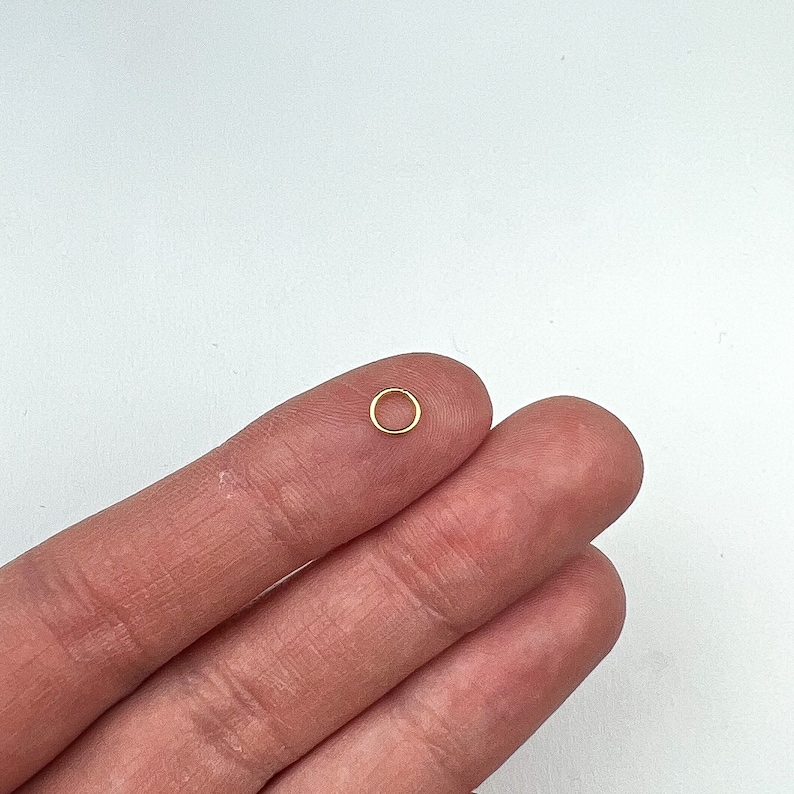 May include: A small, gold-colored jump ring, a type of jewelry finding, is held between two fingers.