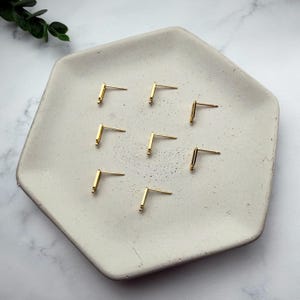 Pode incluir: Sete brincos de pino em metal dourado, cada um apresentando um design simples de barra em forma de L com um pino reto que se estende a partir do canto. Os brincos est&atilde;o dispostos num padr&atilde;o disperso sobre uma bandeja hexagonal clara.