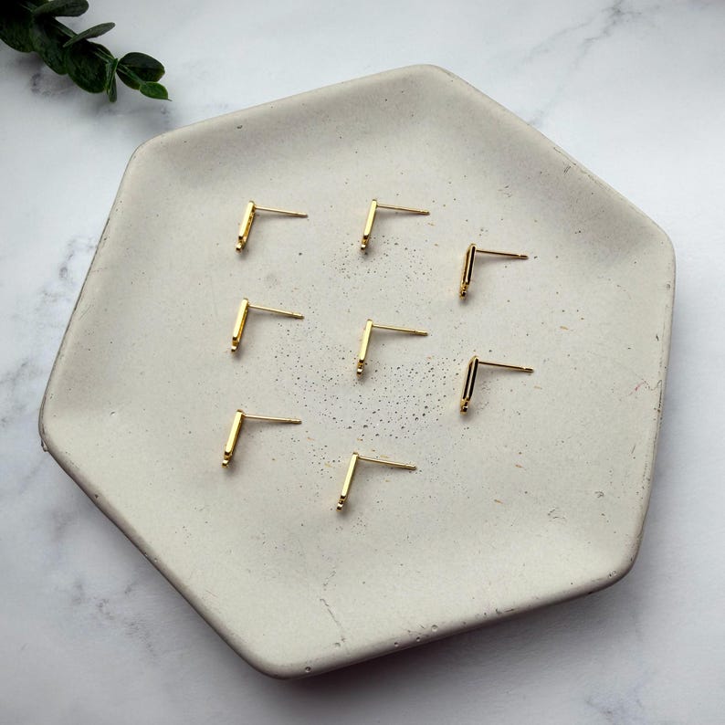 Pode incluir: Sete brincos de pino em metal dourado, cada um apresentando um design simples de barra em forma de L com um pino reto que se estende a partir do canto. Os brincos est&atilde;o dispostos num padr&atilde;o disperso sobre uma bandeja hexagonal clara.