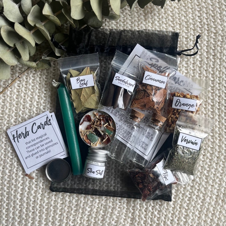 May include: A black mesh bag filled with small bags of herbs and spices, including bay leaf, sandalwood incense, cinnamon, orange peel, vervain, and rose hips. There is also a green candle, a small jar of sea salt, and a card that says "Herb Cards!"
