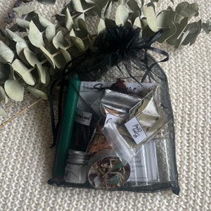 May include: A black mesh bag filled with various herbs, spices, and a green candle. The bag also contains a small jar of oil and a small bag labeled "Bay Leaf".