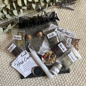 May include: A black mesh bag filled with various herbs and ingredients for spell jars. The bag contains small glass jars filled with dried herbs, a white candle, a small tin of sea salt, and a stack of cards labeled "Herb Cards". The cards list magical correspondences for different herbs.