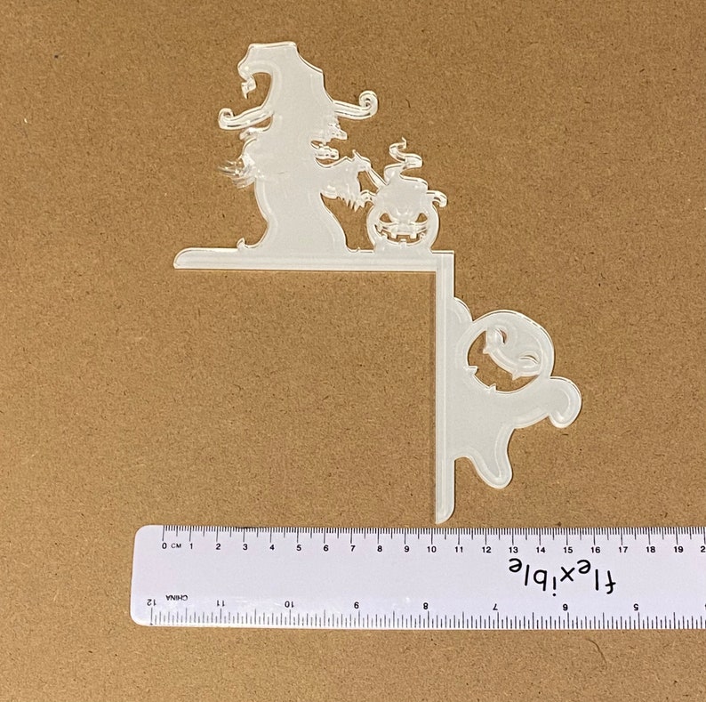 May include: A clear acrylic Halloween corner decoration. The design features a witch with a pumpkin and a ghost. The ruler shows the decoration is approximately 18 cm wide. Ideal for adding a spooky touch to any Halloween display.