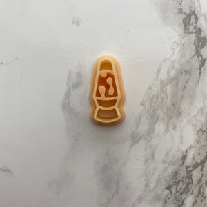 Puede incluir: Un cortador de galletas de plástico blanco con forma de lámpara de lava. El cortador tiene una parte superior redondeada y una base estrecha. El diseño de la lámpara de lava incluye una bombilla en la parte superior y un diseño ondulado y abstracto en el medio.