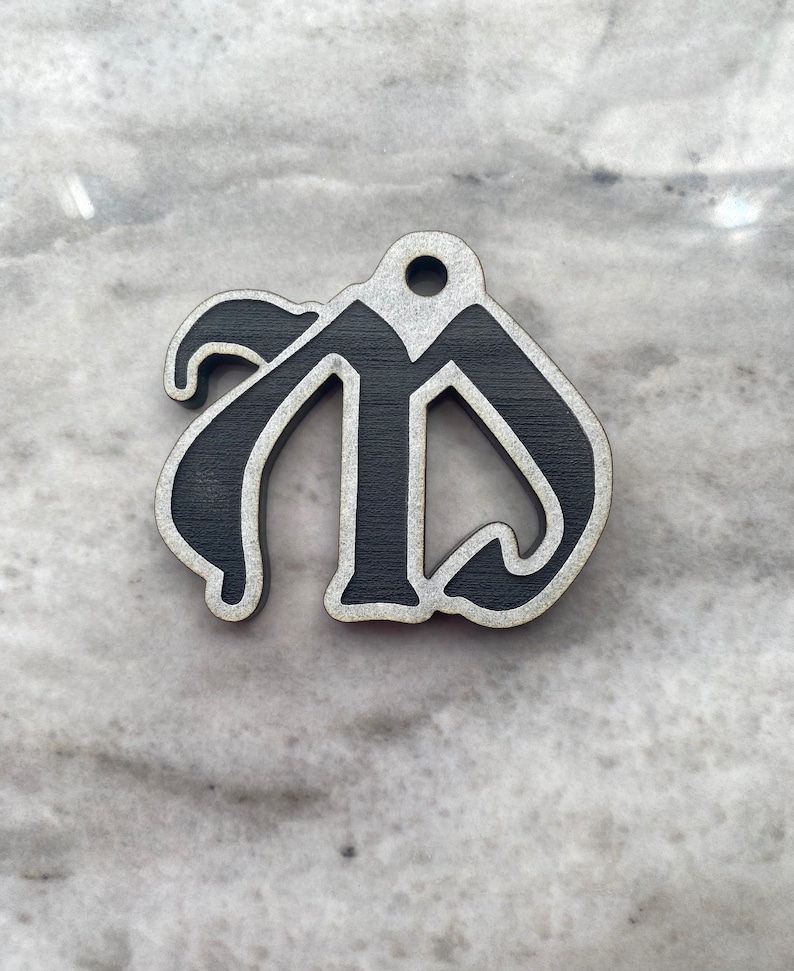 May include: A black wooden ornament shaped like the letter "M" in a stylized font. The ornament has a hole at the top for hanging.