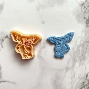 Pode incluir: Um cortador de biscoitos de plástico bege claro em forma de Gremlin, ao lado de um objeto azul em forma de Gremlin. O cortador de biscoitos tem traços faciais e forma corporal detalhados. O objeto azul tem uma superfície lisa e brilhante.