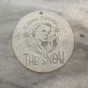 Könnte beinhalten: Ein runder Holzanhänger mit weißem Hintergrund und einem Schwarzweißbild einer maskierten Figur, die ein Messer hält. Der Text "Slashing Through The Snow" ist um das Bild herum geschrieben.