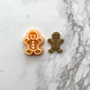 May include: A gingerbread man cookie cutter and a brown gingerbread man cookie. The cookie cutter is made of plastic and has a smiling face, buttons, and arms. The cookie is made of gingerbread and has a smiling face, buttons, and arms.