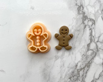 May include: A gingerbread man cookie cutter and a brown gingerbread man cookie. The cookie cutter is made of plastic and has a smiling face, buttons, and arms. The cookie is made of gingerbread and has a smiling face, buttons, and arms.